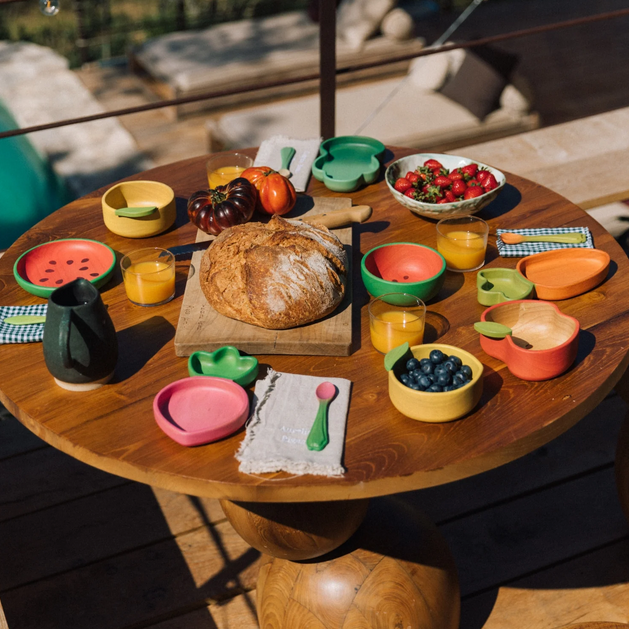 Wooden table with colorful wooden plates, bowls, and a loaf of bread on an outdoor patio.