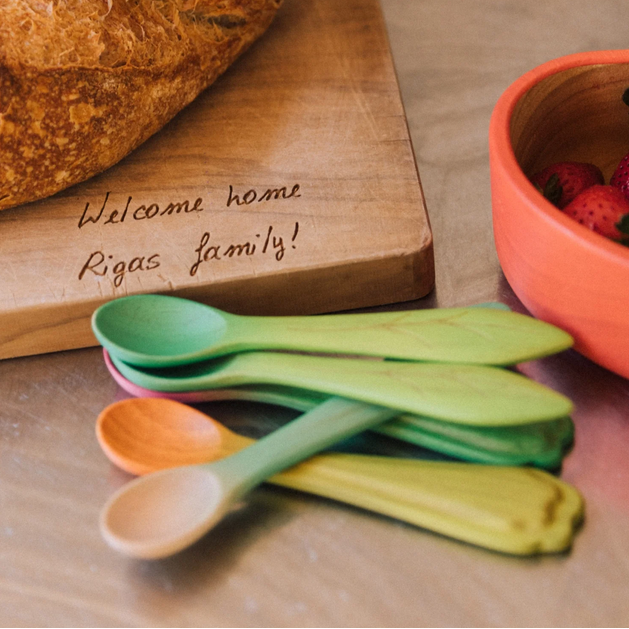 Colorful wooden spoons on a wooden surface with a bowl of strawberries and a cutting board with a message.