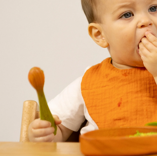 Baby in an orange bib eating with a spoon and showing food on hands
