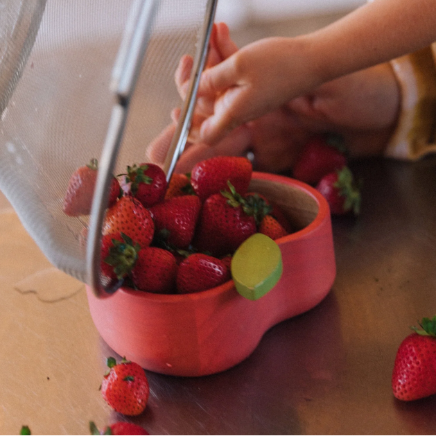 Child's hand reaching into a pink bowl filled with strawberries on a wooden surface.