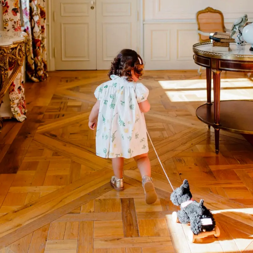 Child in a formal dress walking a toy dog on a wooden floor in an elegant room.