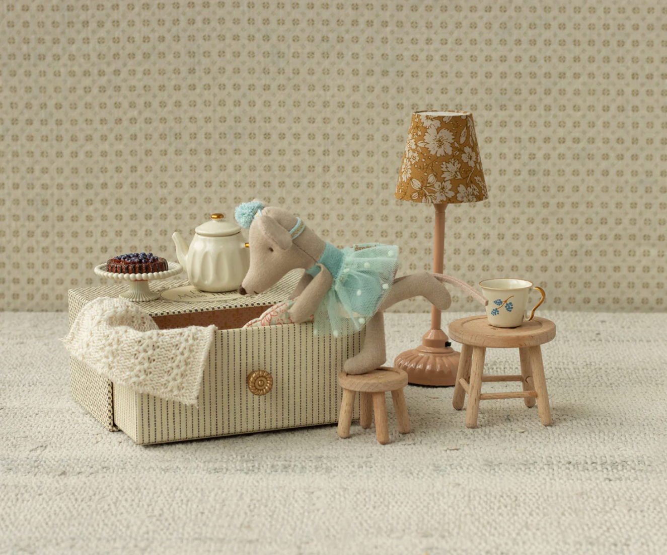 Miniature tea set with accessories on a textured surface with patterned wallpaper.