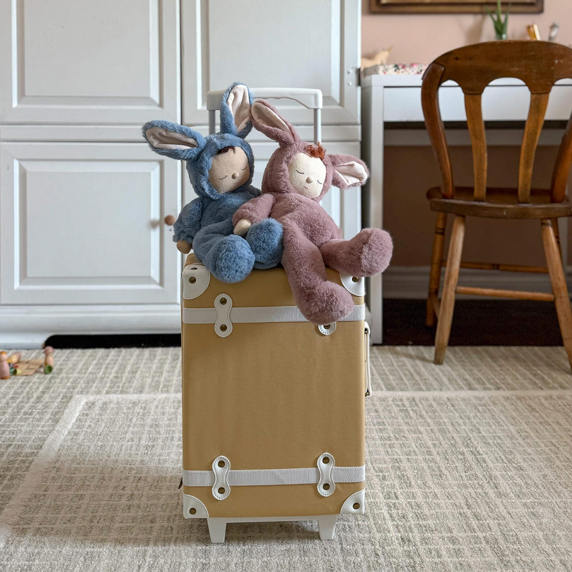 Two stuffed animal bunnies on a beige suitcase in a room with a wooden chair and white cabinets.
