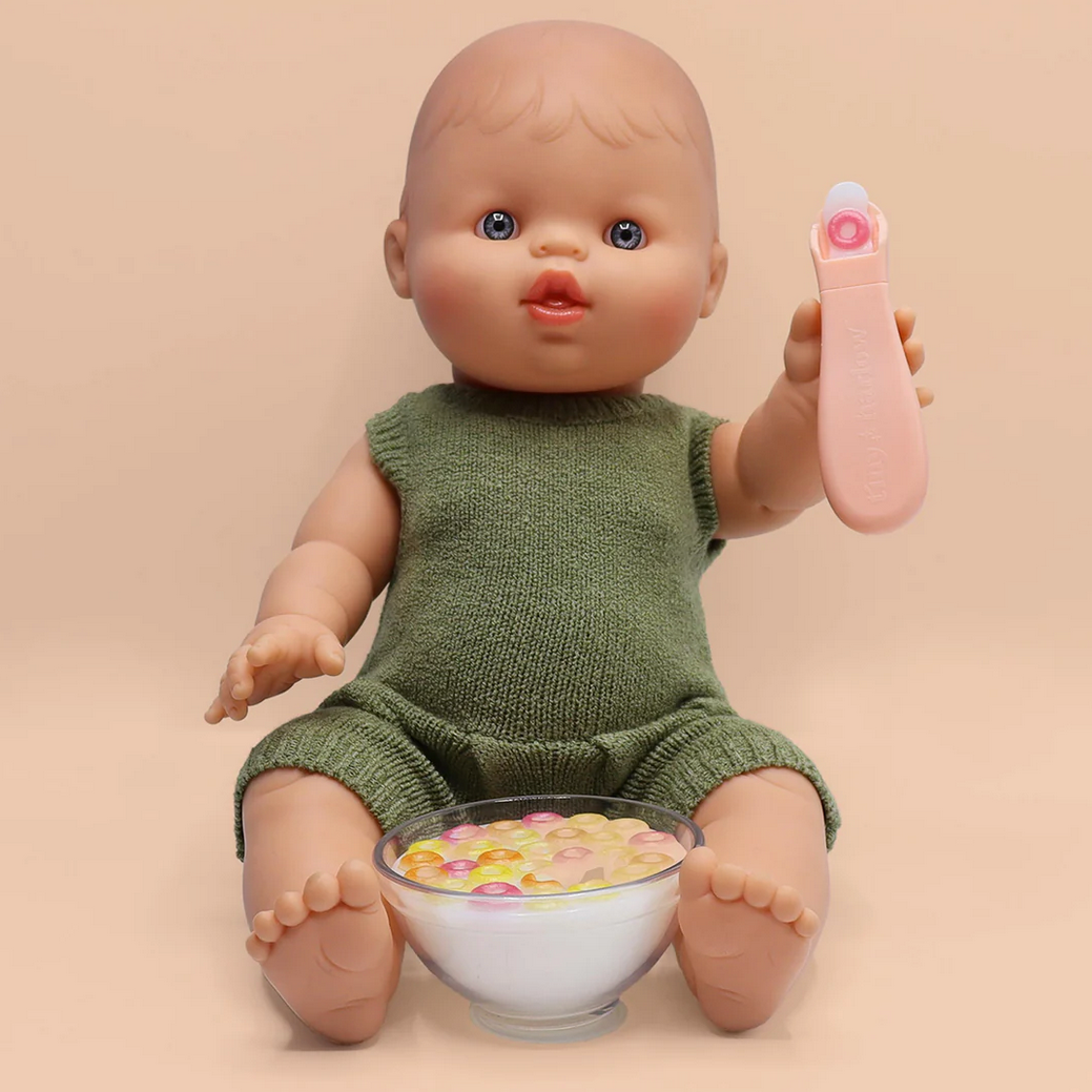 Baby doll in a green outfit holding a pink spoon with a bowl of cereal on a beige background