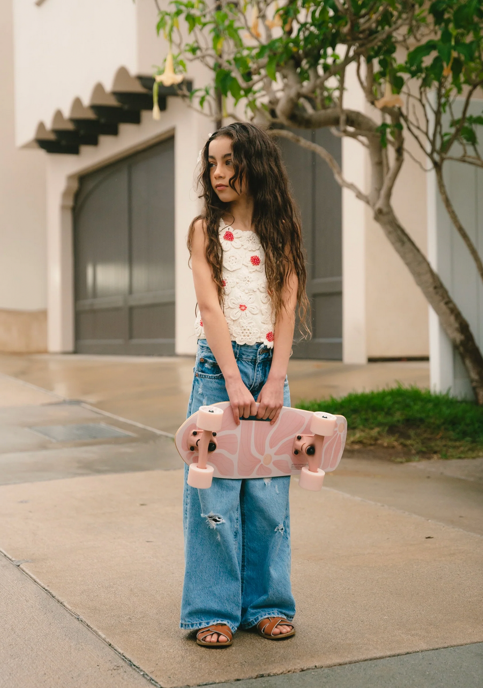 Young girl holding a pink skateboard outdoors on a sidewalk.