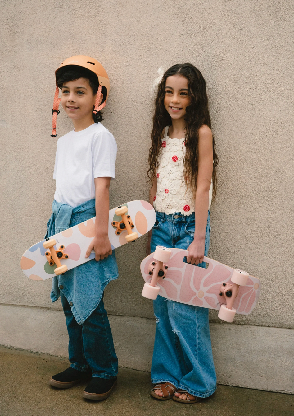 Two children holding small skateboards against a plain wall.