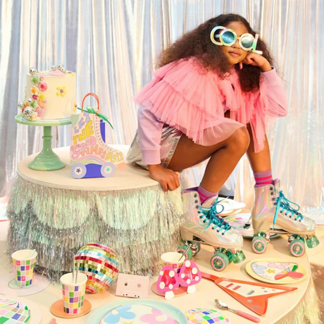Child in pink outfit with roller skates sitting at a decorated table with birthday cake and toys.