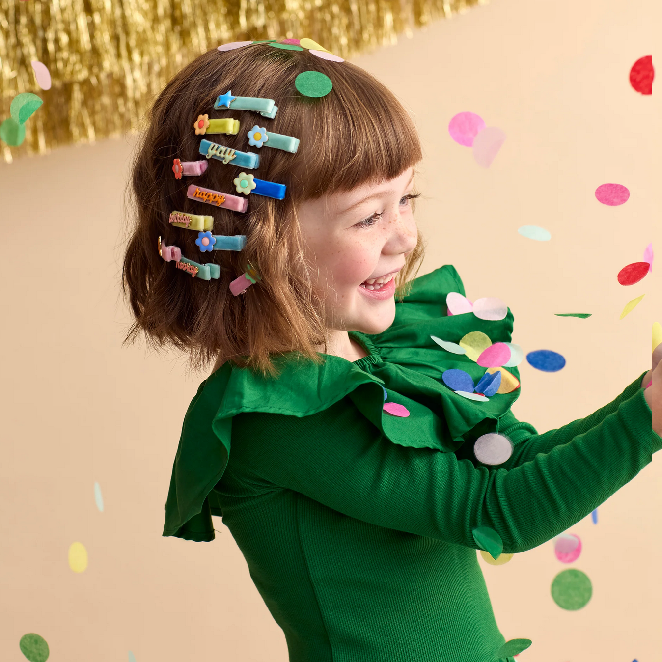 Child in a green dress with colorful hair clips on a beige background with confetti