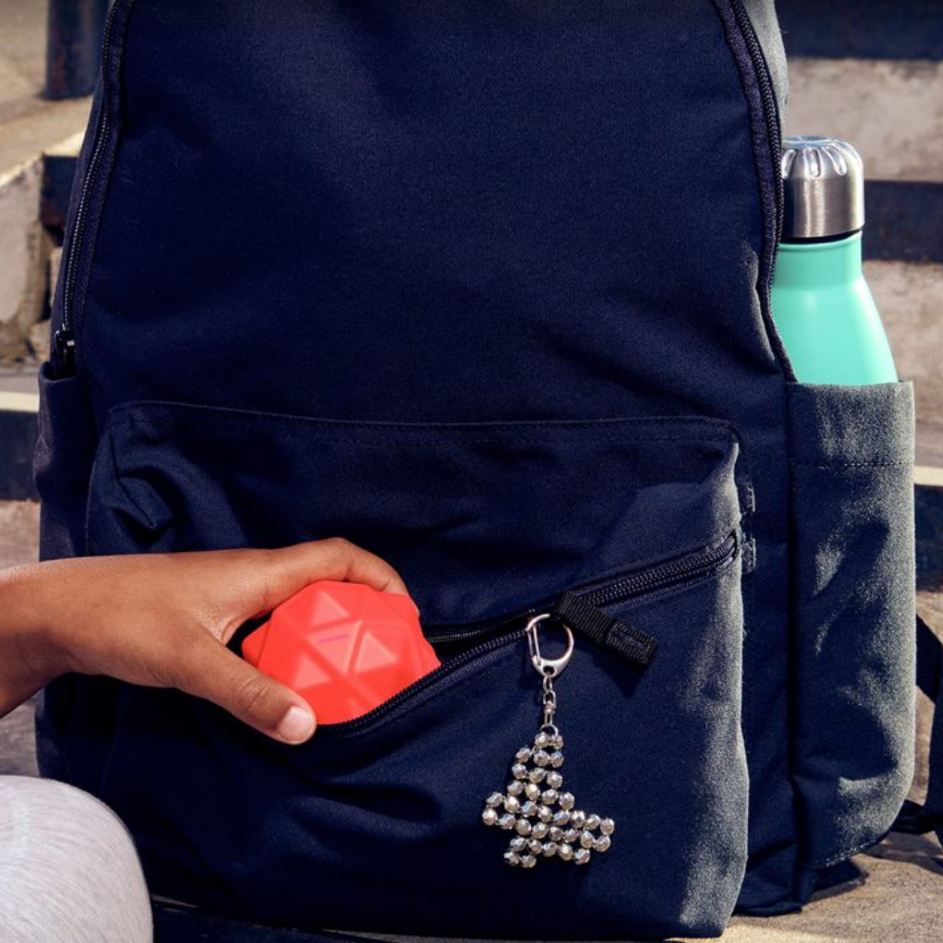 Person holding a red bouncy ball next to a blue backpack with a silver pendant.