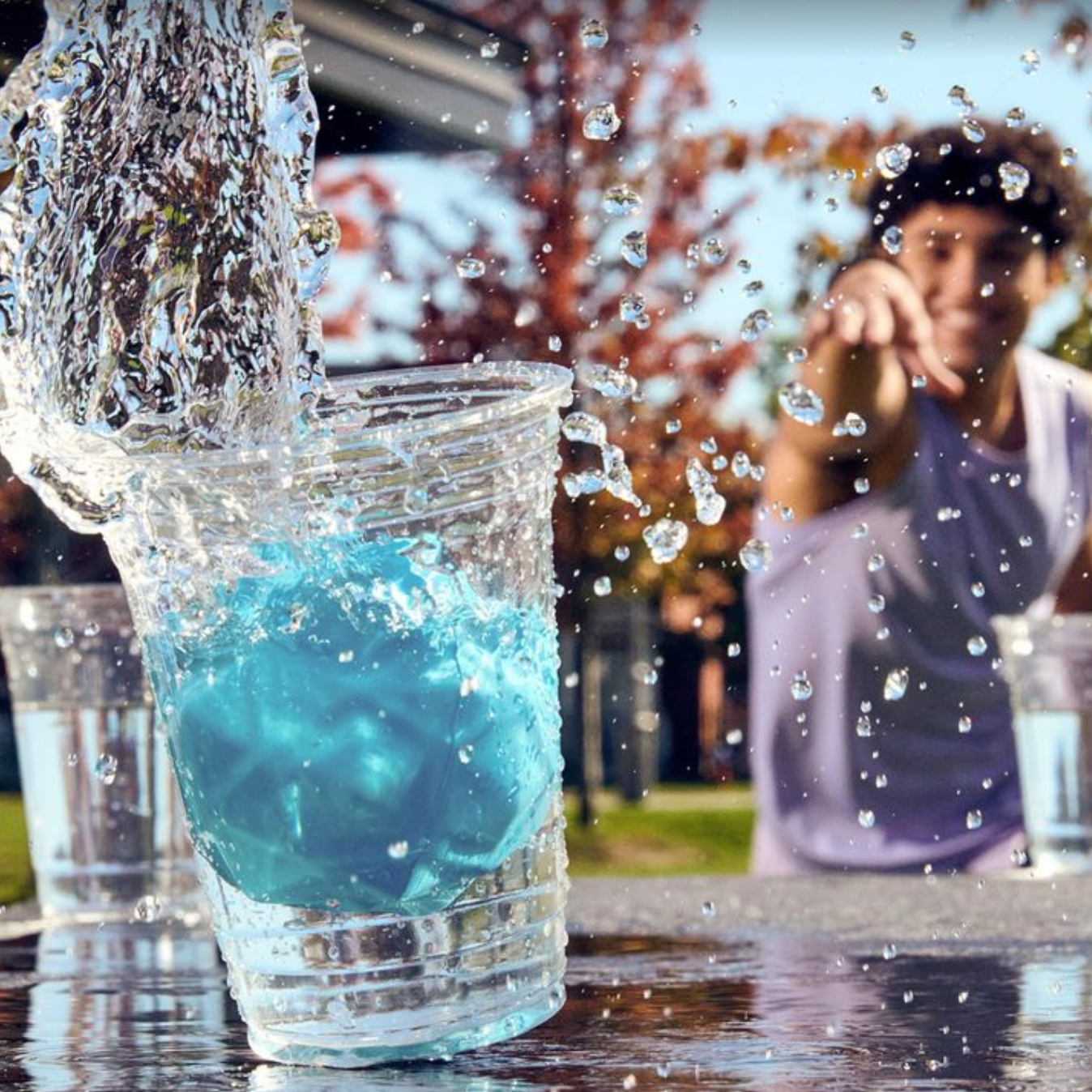 Blue liquid being poured into a clear cup with a blurred background of people playing outdoors.