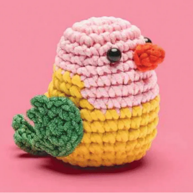 Crocheted bird toy with pink head, yellow body, green feet, and orange beak on a pink background