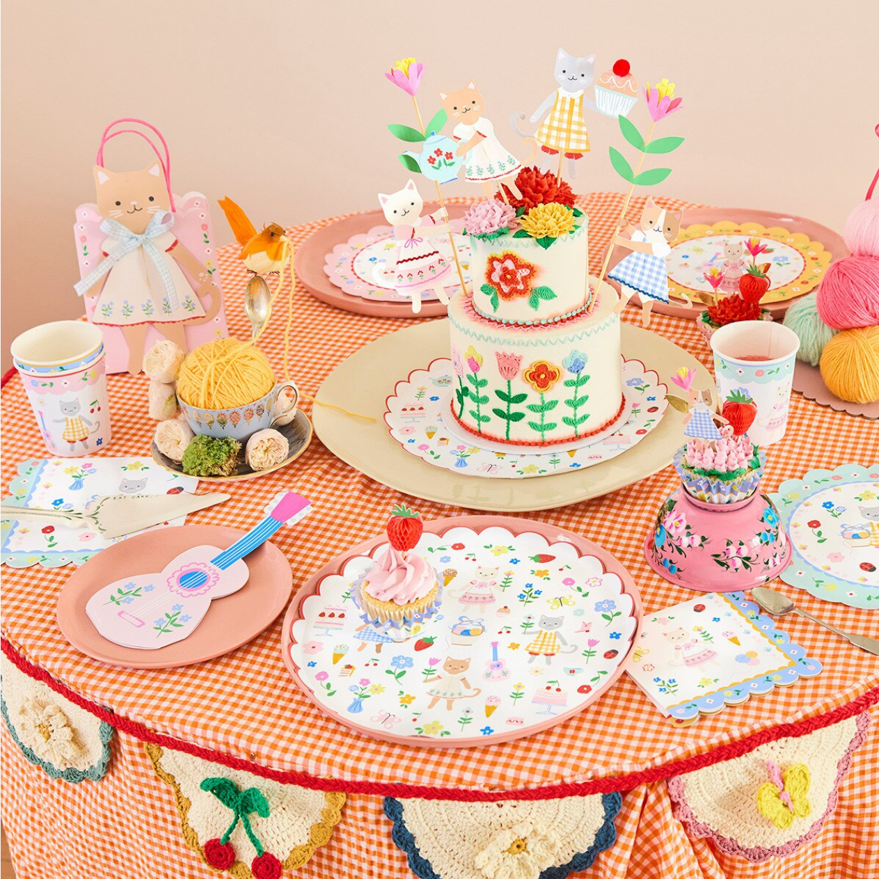 Children's play table with toy cakes, dolls, and decorative items on a checkered tablecloth.
