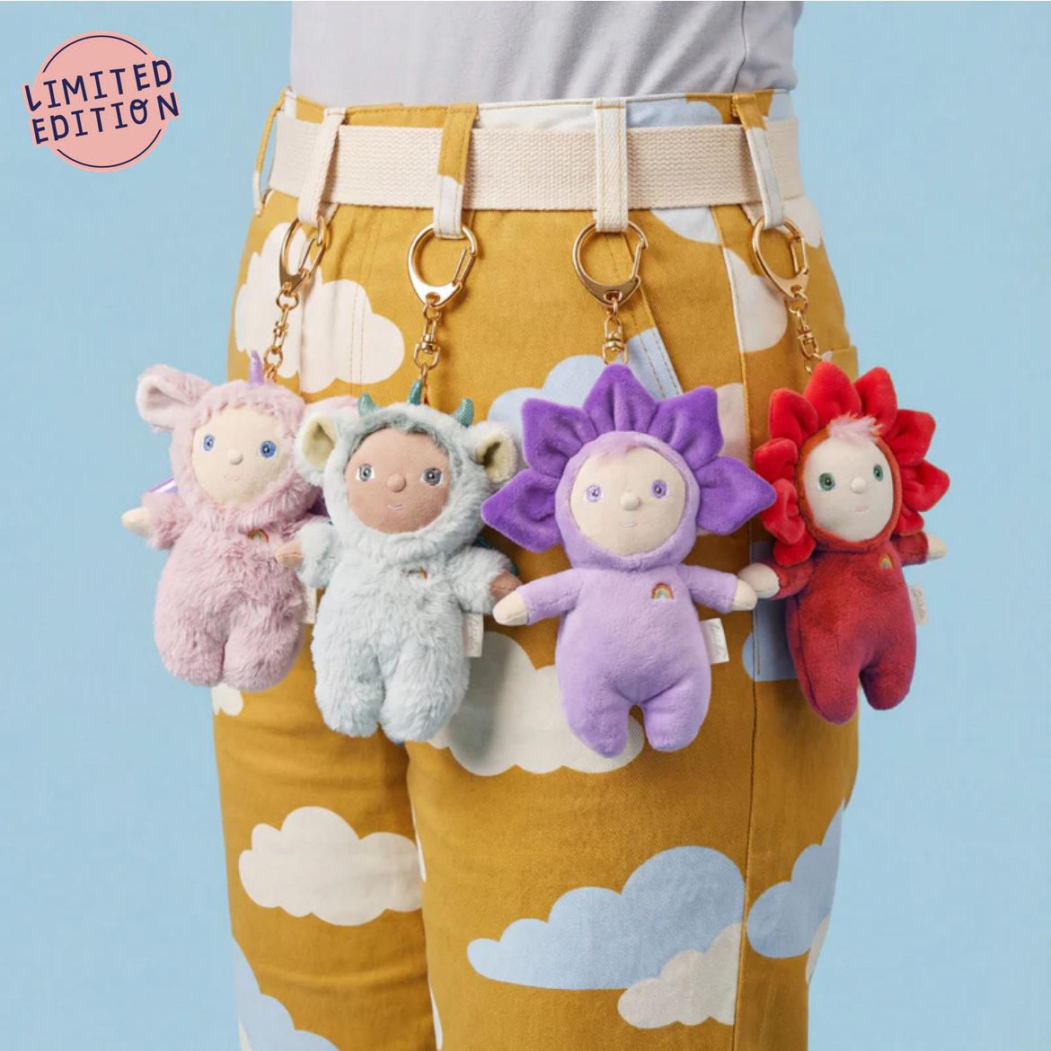 Four colorful plush keychains hanging from a belt on mustard yellow pants with cloud pattern against a light blue background.