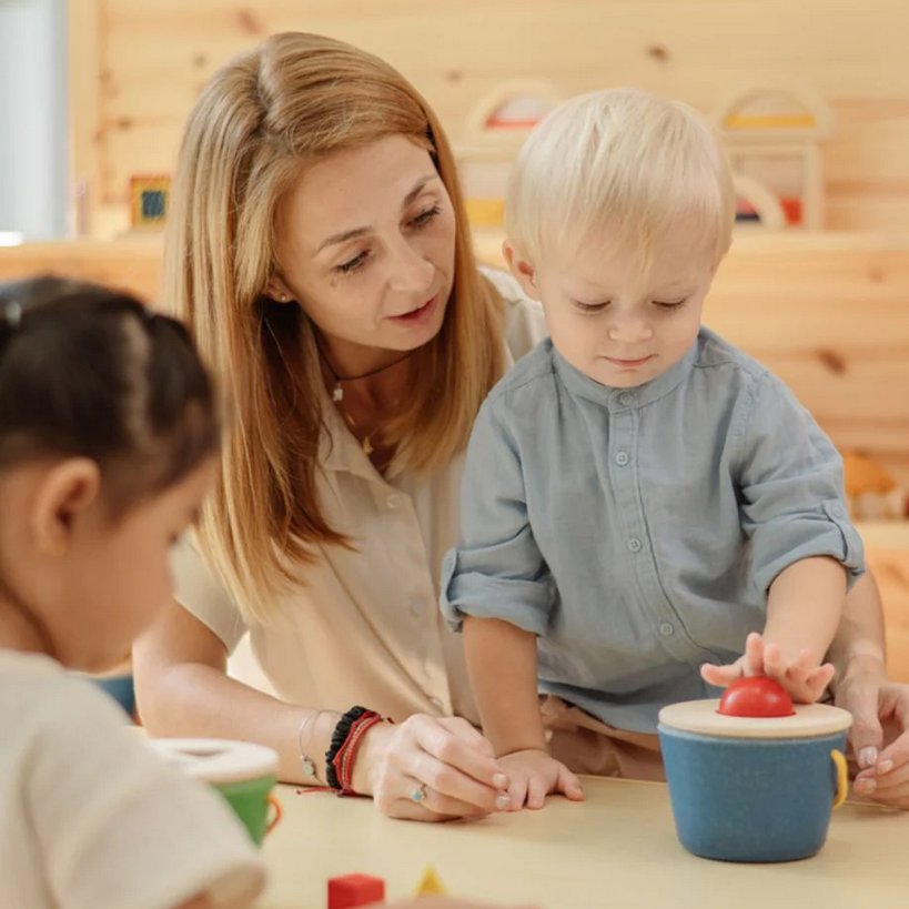 Woman and two children playing with a toy at a table in a classroom setting.