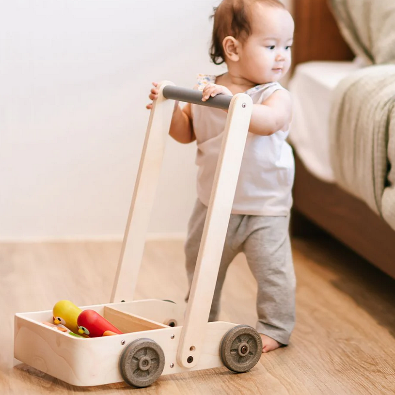 Child pushing a wooden toy cart with wheels on a wooden floor.