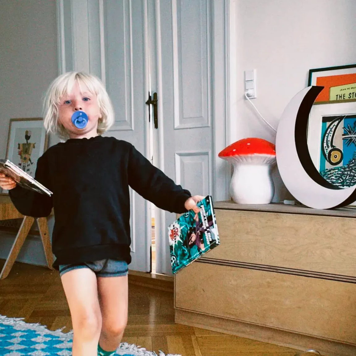 Child holding a book and a toy in a room with a wooden dresser and decorative items.