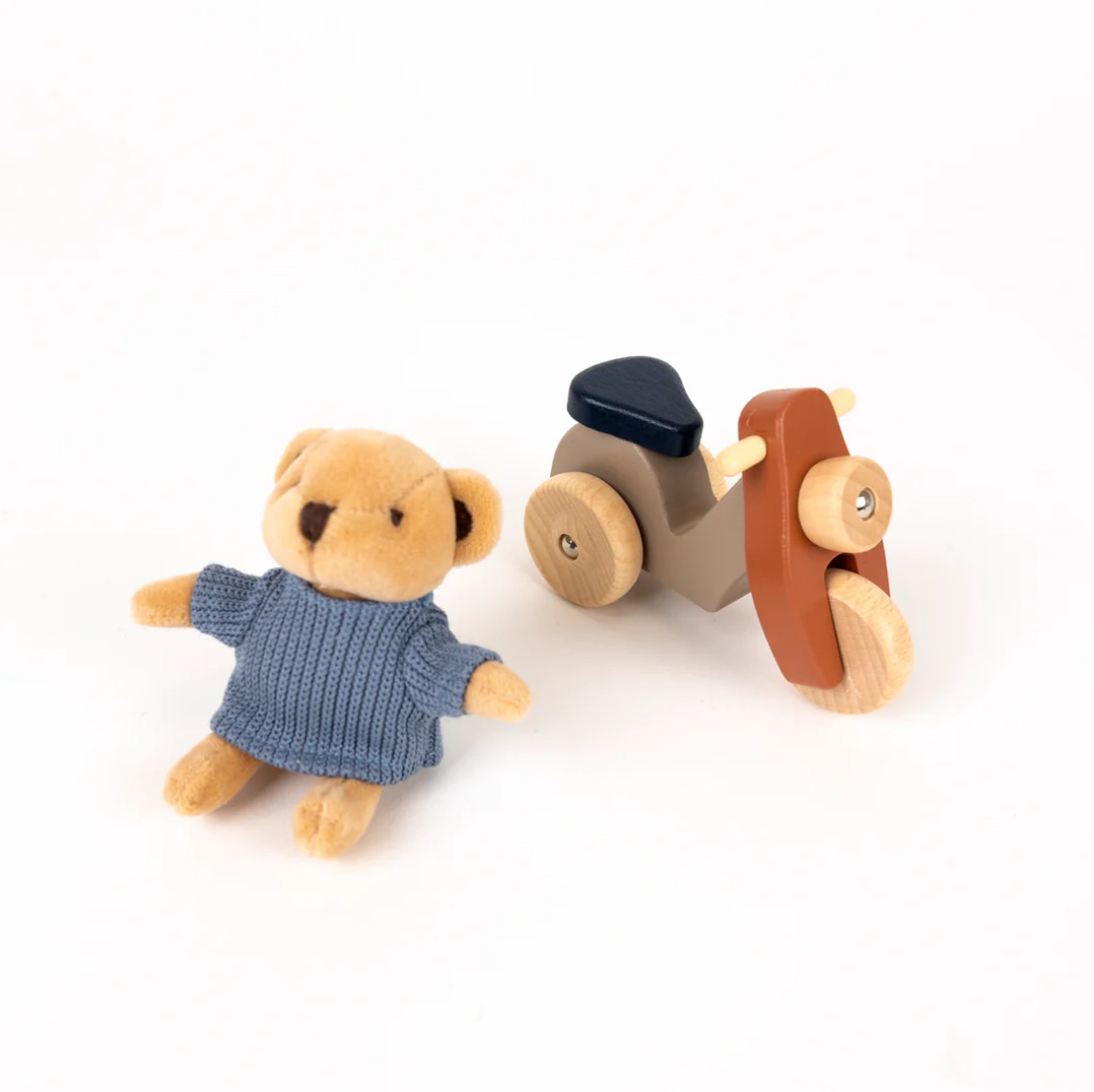 Small teddy bear in a blue dress next to a wooden toy vehicle on a white background