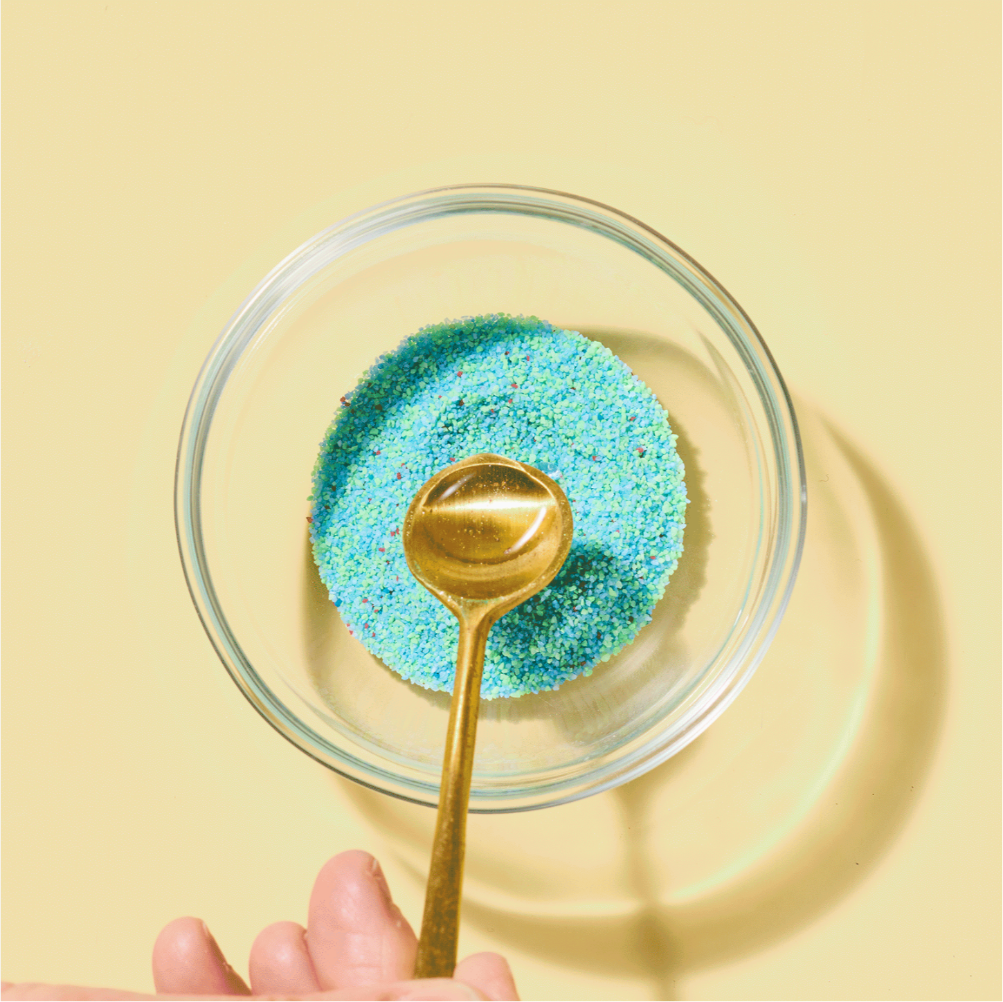 Glass bowl with blue glittery sand and a gold spoon on a yellow background