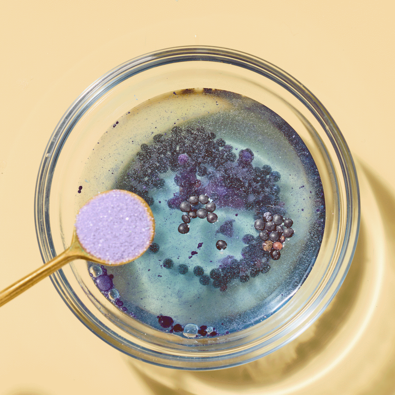 Glass bowl with purple bath salts and a gold spoon on a yellow background