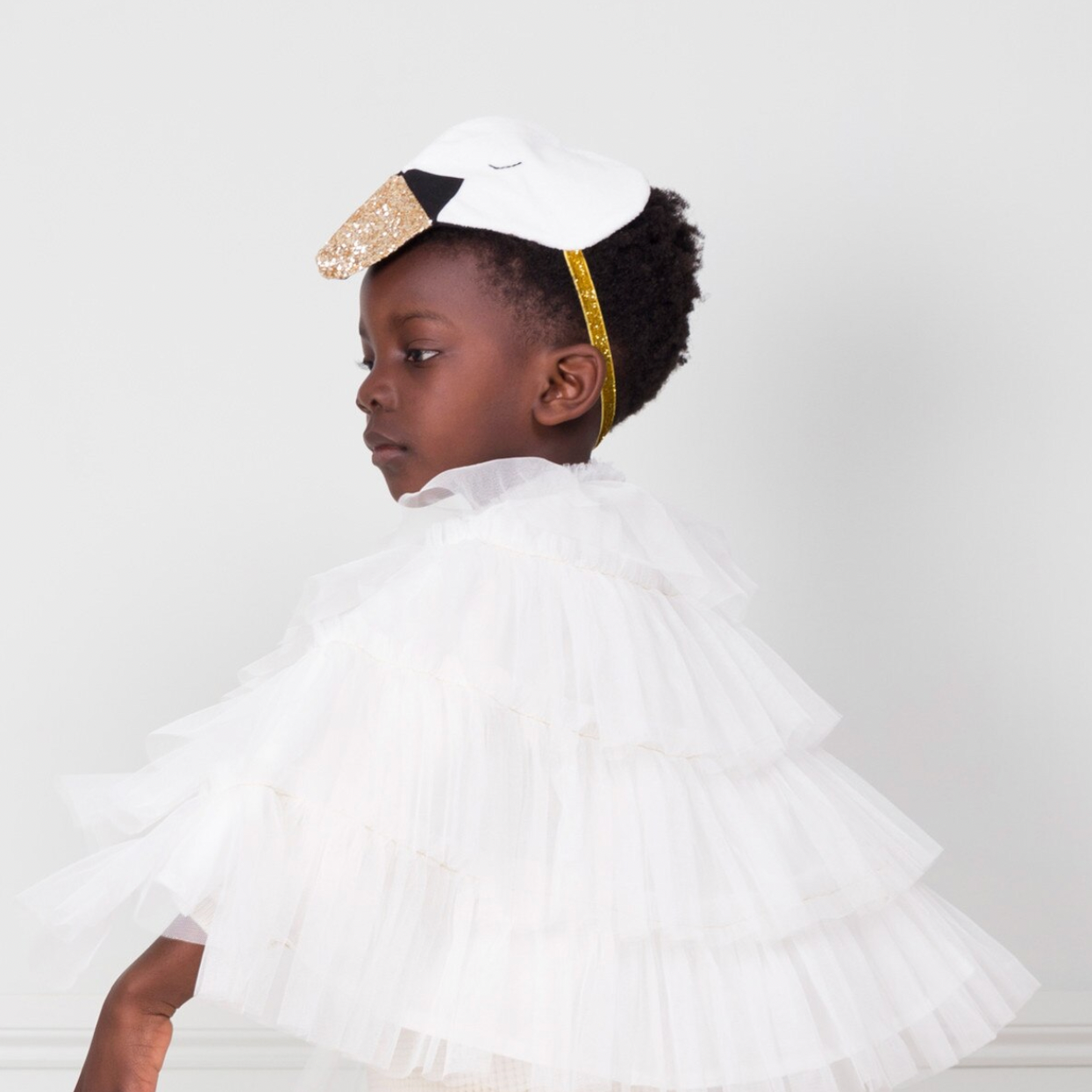 A young child wearing a white tiered tulle dress with a white bow headband featuring a gold glitter accent, shown in profile against a light gray background.