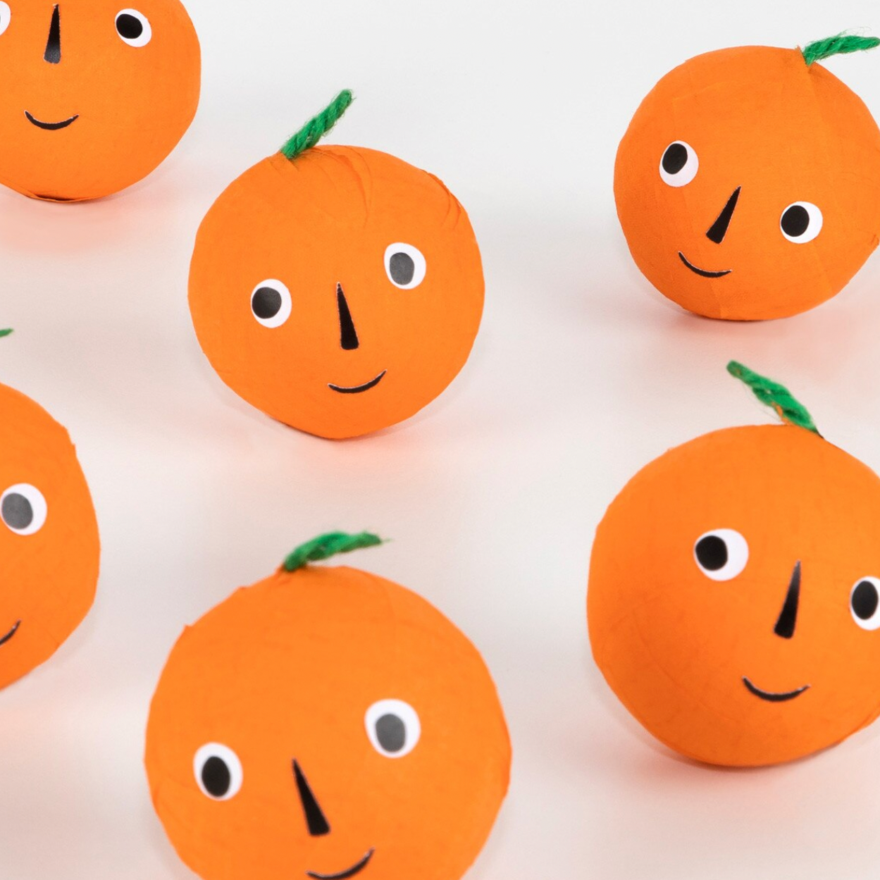 A set of handmade orange paper decorations with simple paper faces and green stems, arranged on a light background.