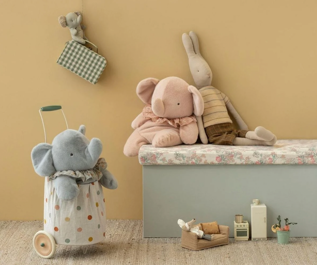 A styled nursery scene with soft plush toys and toys arranged on a bench against a warm beige wall. A large pink elephant plush sits on the bench, a gray rabbit plush leans behind it, and a gray elephant plush is inside a white polka-dot laundry bag stroller on the floor.