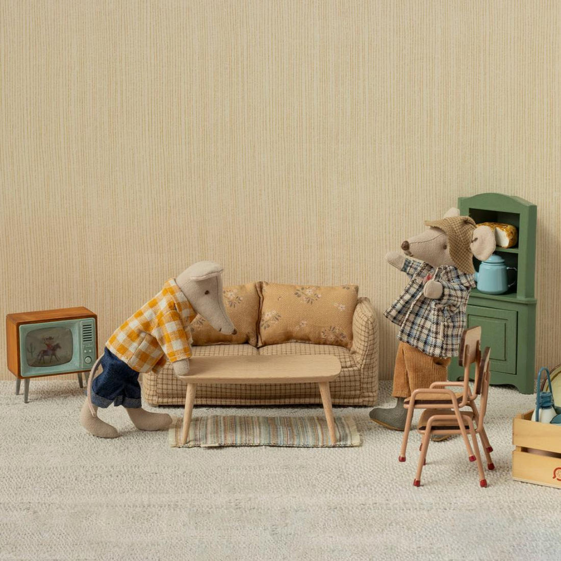 A miniature dollhouse interior scene with two animal figurines posed around a small sofa, table, chairs, cabinet, TV, and storage crate against a beige textured wall.
