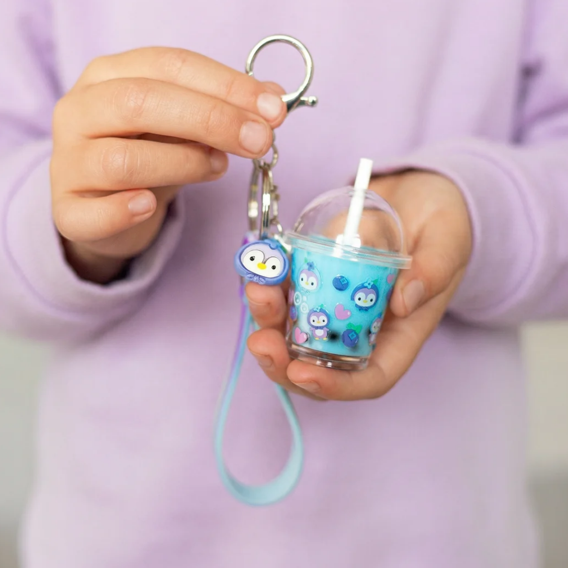 A person in a lavender sweatshirt holds a keychain featuring a small blue cup with a straw, cartoon owl characters, and a light blue wrist strap.