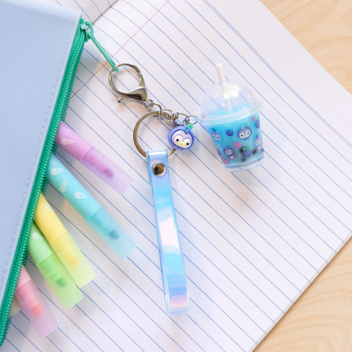 A pastel-colored keychain set displayed next to a blue zippered pencil case on an open lined notebook. The keychain features a blue penguin charm, a matching blue penguin drink cup accessory with a straw, and a translucent blue-and-white striped wrist strap attached to silver key rings.