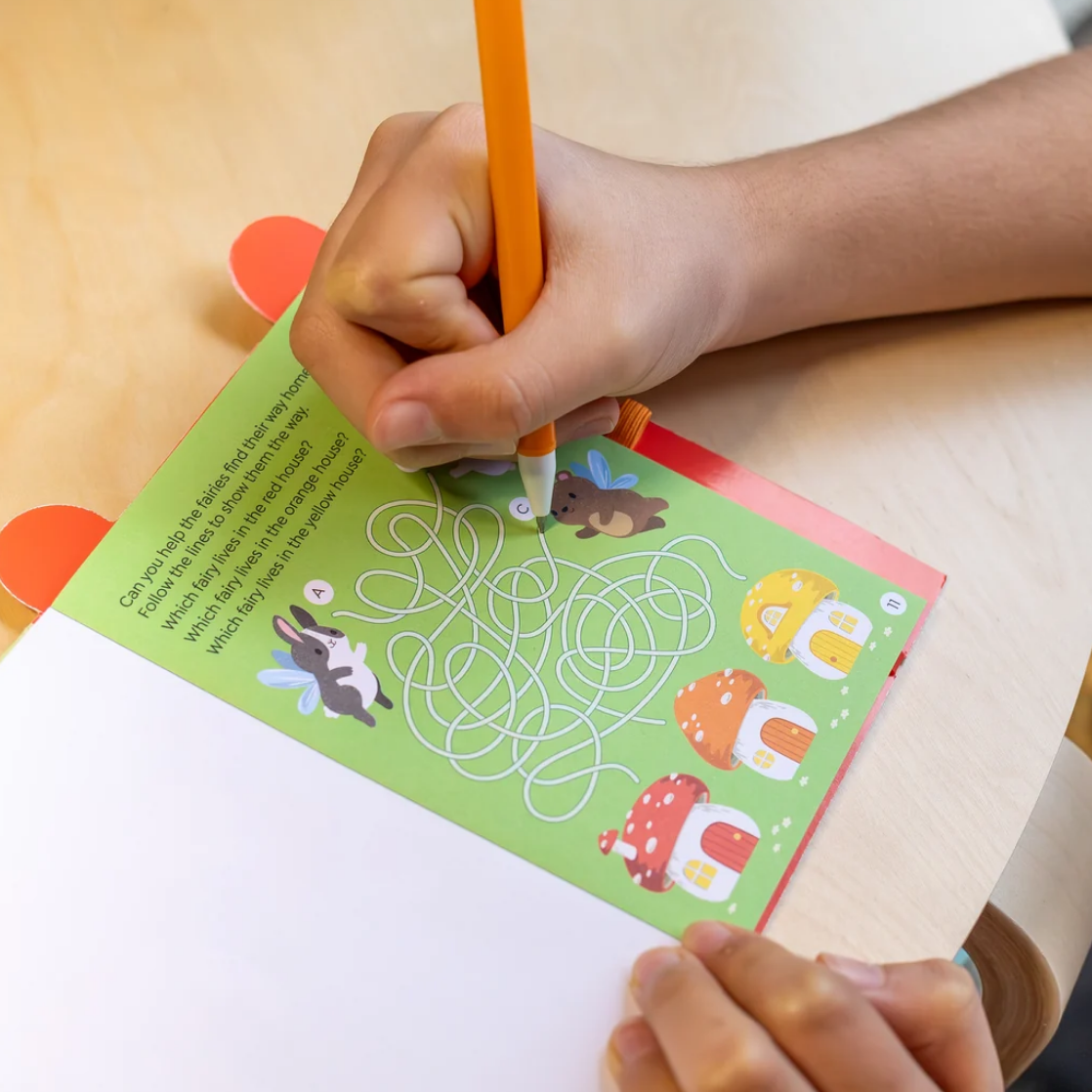 A child's hand holding an orange pencil as they trace a matching game page on a wooden desk. The page is green with cartoon animals and colorful houses.