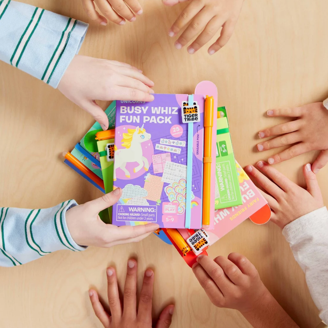 A children's desk activity set called 'Busy Whiz Fun Pack' by Tiger Woods is held by multiple children's hands on a light wood table. The purple box features a unicorn illustration, and an orange pen is tucked into the top of the stack of colorful activity sheets.