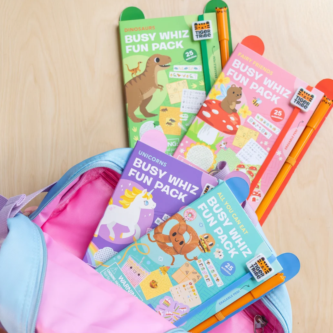 Four colorful 'Busy Whiz Fun Pack' activity book sets fanned out inside a pastel pink and blue backpack on a light wood surface. The packs feature dinosaur, unicorn, mushroom, and bear illustrations, and are styled with orange and blue pens.