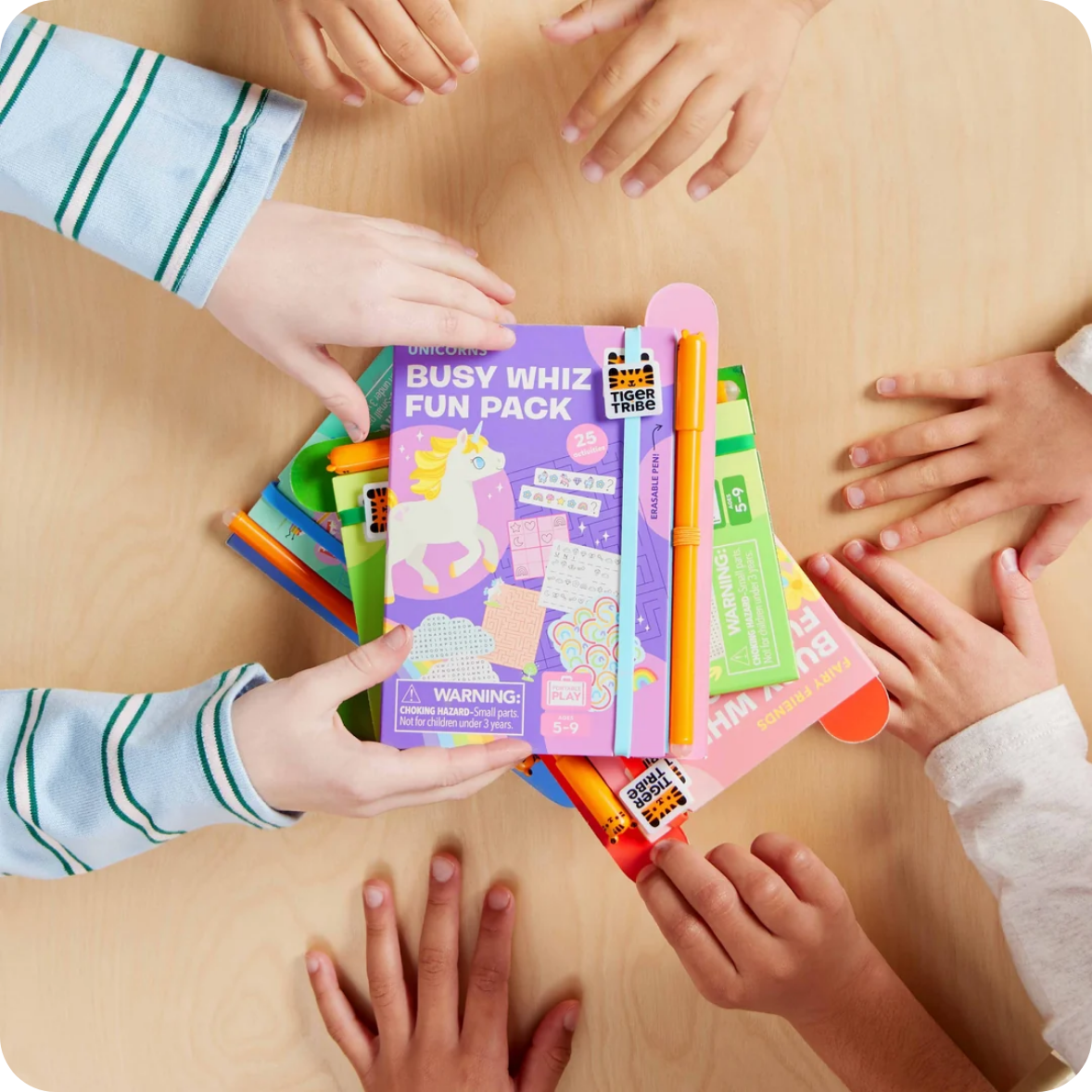 A children's desk activity set called 'Busy Whiz Fun Pack' by Tiger Woods is held by multiple children's hands on a light wood table. The purple box features a unicorn illustration, and an orange pen is tucked into the top of the stack of colorful activity sheets.