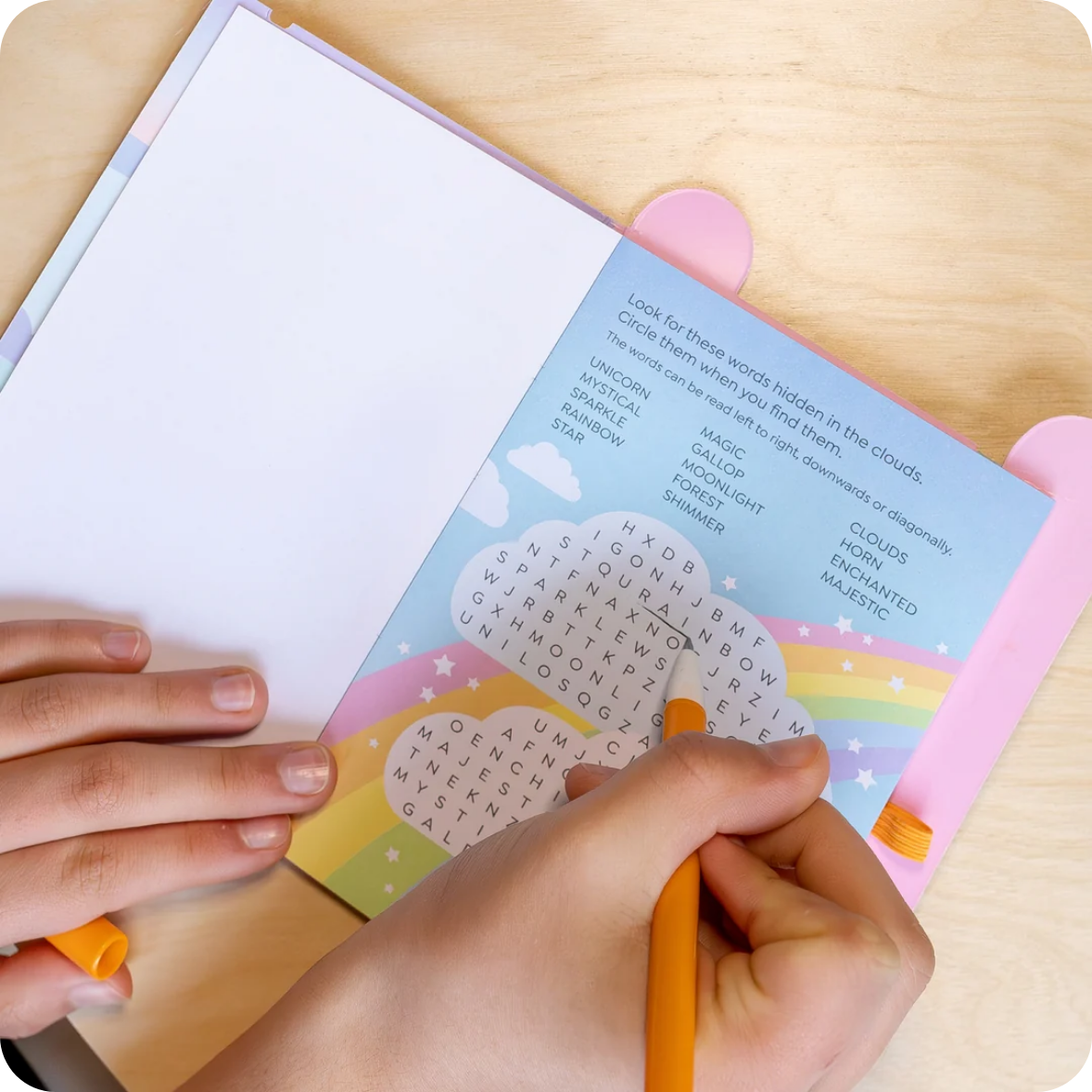 A close-up of hands using a colorful cloud-word activity book on a light wood tabletop. One hand holds an orange pen over a pastel rainbow and cloud page, while the other hand holds the open book.