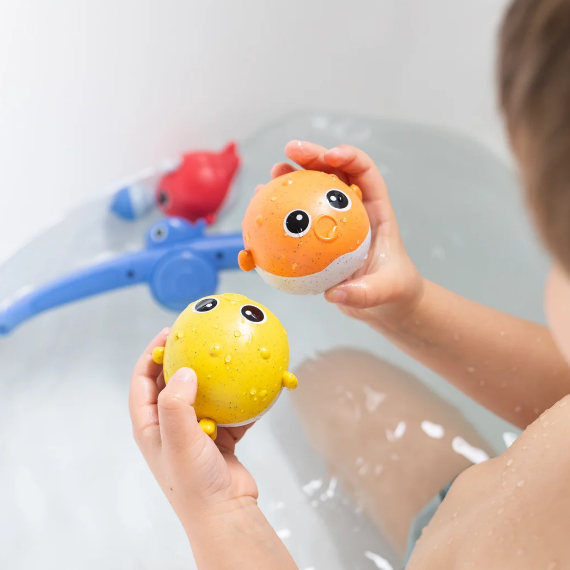 A child holds two small squishy bird-shaped bath toys, one orange and one yellow, above a bathtub with water and other bath toys visible in the background.