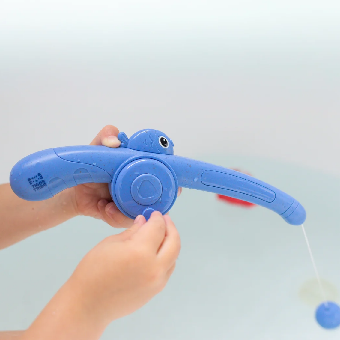 A blue plastic fish-shaped bath toy being held by a child's hands in a bathtub. The toy has a curved fishing rod design with a small fish head on the handle and a small pull string dangling from the tip.