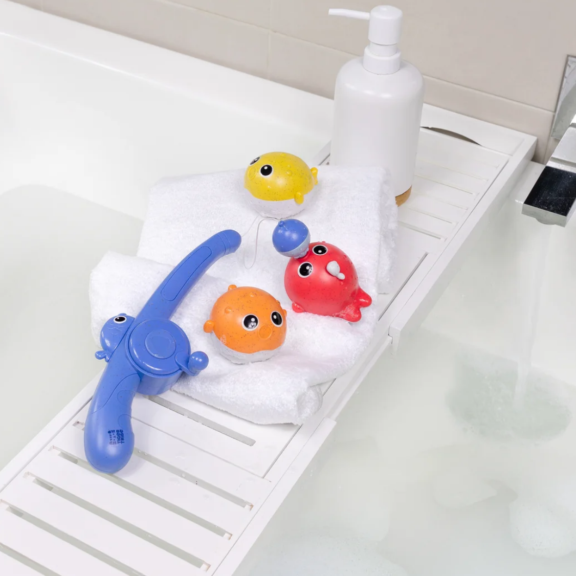 A set of colorful baby bath toys arranged on a white bathtub caddy next to a white pump bottle, with water running into the tub.
