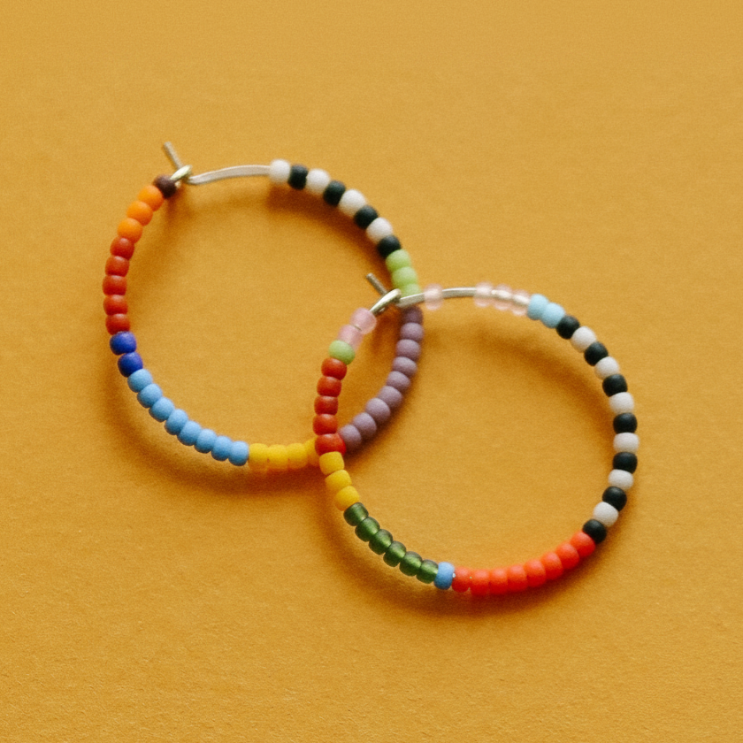 Two handmade rainbow beaded hoop earrings on a textured orange background.