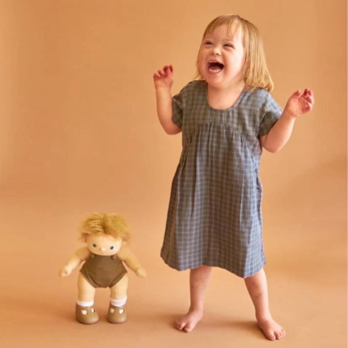 A young child wearing a blue-gray and white plaid short-sleeve dress stands barefoot against a warm tan studio backdrop, smiling and laughing while holding nothing; a small doll stands beside them. The product appears to be the dress, but because a person is present the subject_type is Lifestyle.