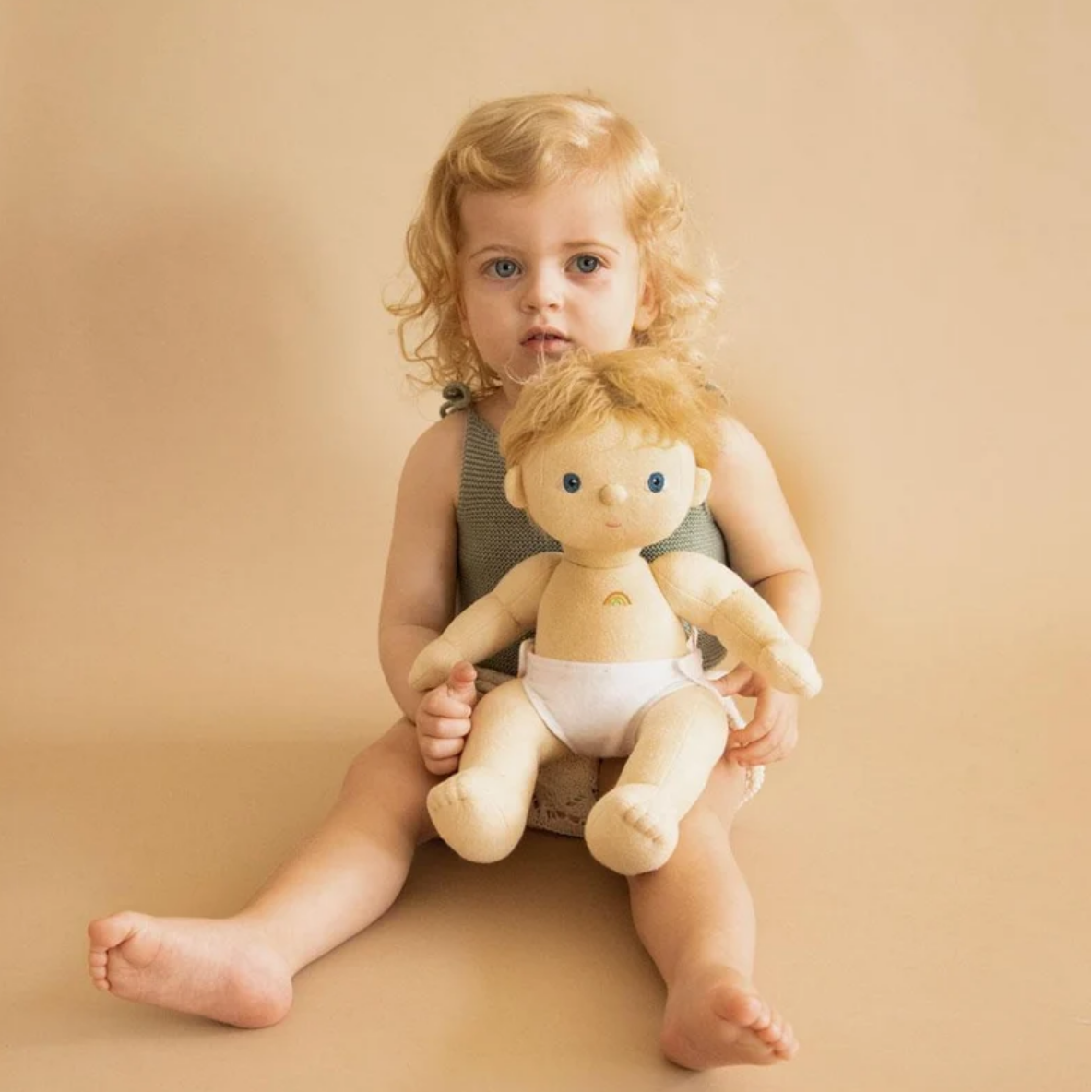 A toddler with curly blonde hair sits barefoot against a warm beige studio background, holding a soft plush baby doll with a white diaper.