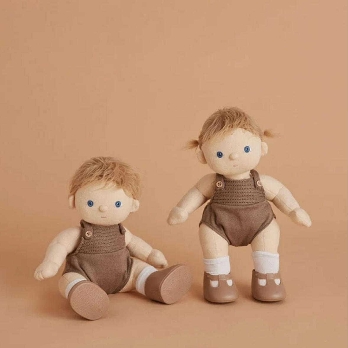 Two soft fabric baby dolls with blonde hair and blue eyes, dressed in brown knit overalls with white socks and brown shoes, posed against a warm beige background.