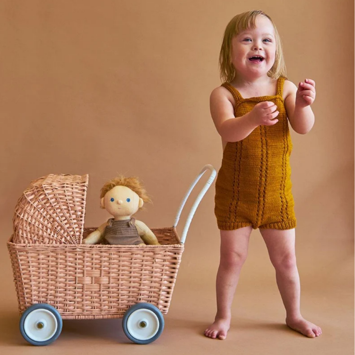 A toddler stands barefoot next to a wicker toy stroller with a doll inside, wearing a mustard yellow knit sleeveless romper, against a warm tan studio backdrop.