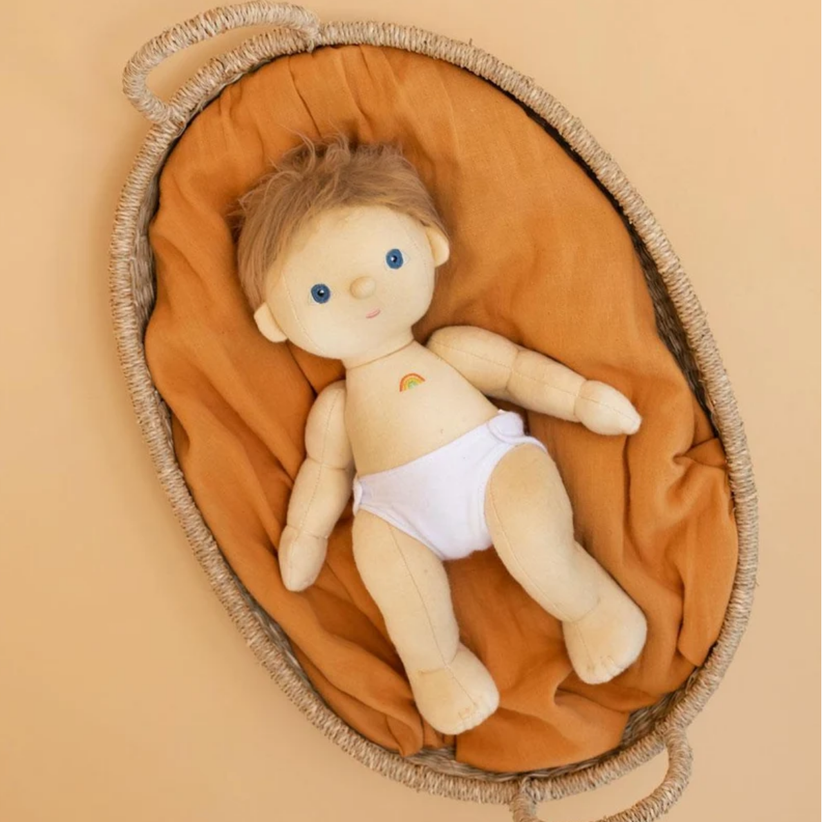 A plush baby doll with light beige skin, short brown hair, blue eyes, and a small rainbow patch on the chest, lying on an orange-brown fabric cushion inside a woven oval basket against a warm beige background.