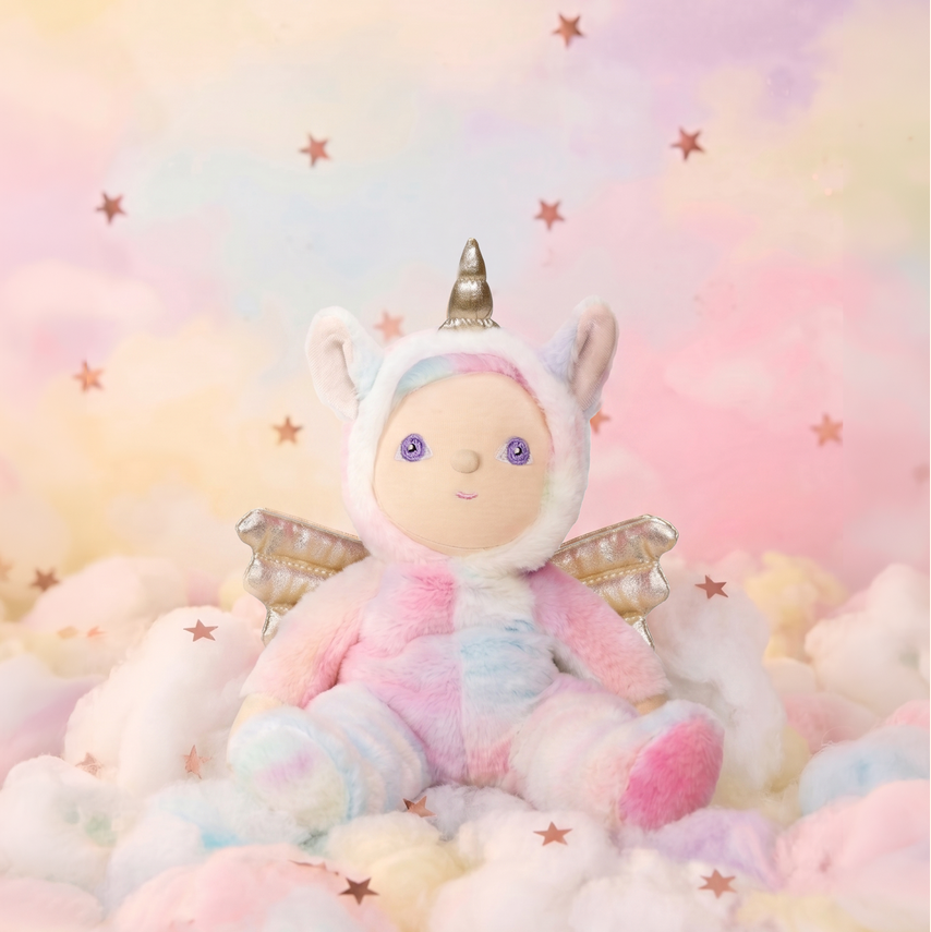 A pastel unicorn plush toy with wings and a horn, sitting on soft clouds against a dreamy pink, yellow, and blue sky backdrop with scattered stars.
