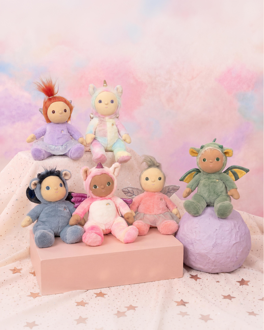 Six pastel-colored plush toys arranged on pink display blocks and a purple ball against a soft pink and blue pastel backdrop. The toys include a red-haired character in purple, a unicorn in mint, a blue bird-like figure, a dark-skinned bunny in pink, a pink fairy, and a green dragon with yellow wings.