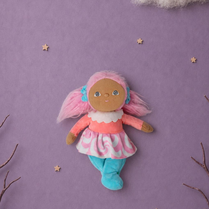A handmade plush doll with pink hair, a pink and white patterned dress, and blue pants, displayed on a purple background with decorative stars, clouds, and twigs.