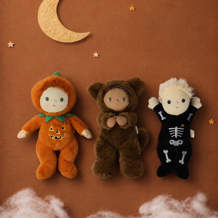 Three plush baby dollies dressed in Halloween costumes are arranged on a warm brown background with a crescent moon, stars, and faux clouds. The left doll is in an orange pumpkin costume, the middle doll is in a brown bear costume, and the right doll is in a black skeleton costume.