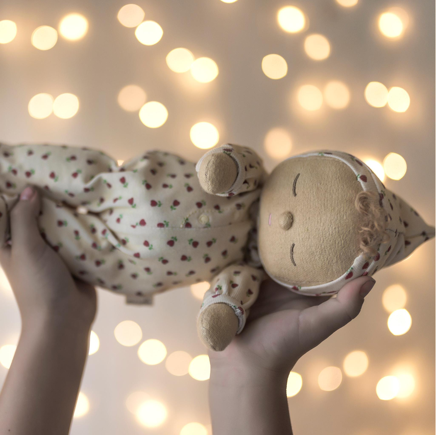 A handmade plush doll with a cream-colored body and red heart patterned clothing, held gently in two hands against a warm bokeh background of glowing lights.