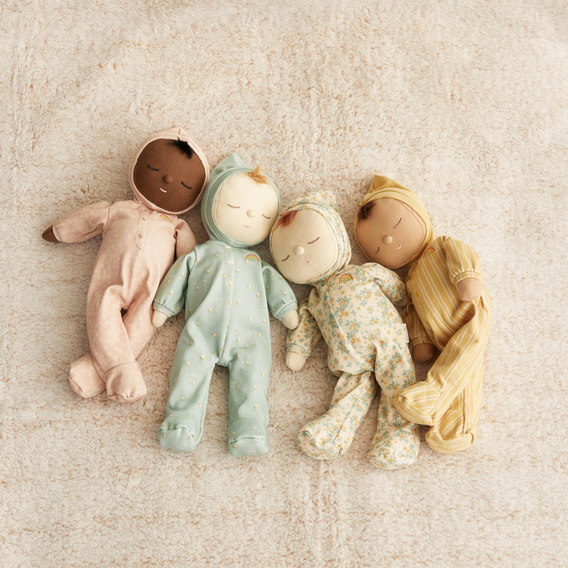 Four handmade fabric dolls dressed in pastel baby outfits, arranged side by side on a textured cream blanket.