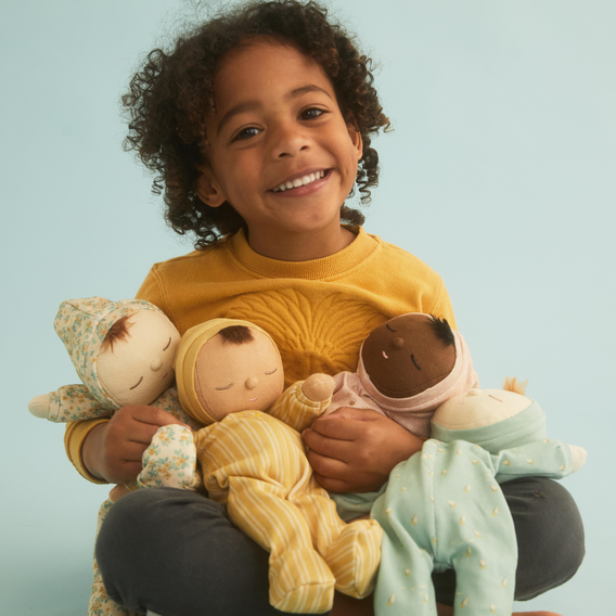 A smiling young child with curly hair sits against a light blue background, holding three soft plush dolls dressed in pastel clothing.